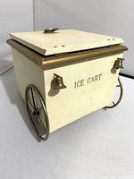 Side view of vintage ice bucket cart showing cream paint, gold wheels and two brass bells, lid partially open