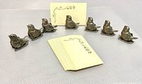 Seven pewter bird place card holders arranged with original place cards in front on textured surface.