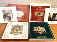 Two White House Christmas ornaments in their respective boxes with brochures displayed behind them, showing detailed designs and packaging.