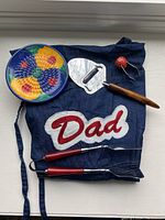 Denim apron with 'Dad' patch, colorful grating plate, berry picker, brown-handled slicer, and two red-handled tongs arranged on a white surface.