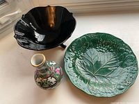 Three items: black ceramic bowl, green decorative plate, and small porcelain vase with floral and gold detail