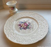 Photo showing the decorative ceramic plate and the bone china vase placed on a windowsill with daylight. The plate has an embossed floral pattern on the rim and a colorful floral cluster in the center. The vase is short, white bone china with floral decals.