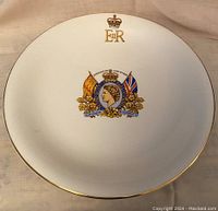 Front of commemorative plate showing Queen Elizabeth II crowned portrait with flags and floral decoration, gold trim along edge, and gold crown with ER initials at top.