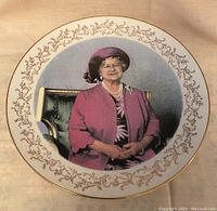 Front of decorative commemorative plate with portrait of Queen Elizabeth, The Queen Mother in purple outfit, gold trim and pattern around edge.