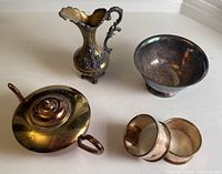 Set of small metal decorative and kitchen items including tea pot, ornate pitcher, napkin rings, and Paul Revere style bowl arranged on a white surface