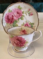 Teacup displayed on clear stand with matching saucer behind, showing floral pattern and gold-trimmed scalloped rims.