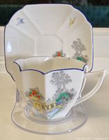 Teacup in front of saucer showing uniquely angled rim and faceted shape with waterside scene design including butterflies and trees.