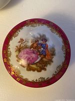 Top view of the domed lid showing the painted scene of two figures in period clothing with burgundy and gold detailing around the edge.