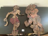 Two fragile antique Indian leather shadow puppets laid flat on a dark surface, highlighting the painted details and wooden rods.