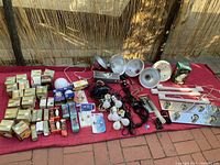 Wide photo showing full lot of assorted vintage light bulbs, fixture reflectors, fluorescent fixtures, electrical cords, and bulbs on red surface outdoors.