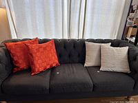 Two orange and two beige/green/blue pillows on a grey tufted couch.
