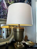 Photograph of a brass table lamp with white fabric shade and an octagonal decorative mirror behind it. Also visible are a small dark figurine and a perforated brass bowl on the table.