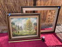 Photo showing signed vintage landscape oil painting with wood canvas frame placed on red fabric background against bamboo fence.