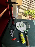 Schwinn bike pump, Prince tennis racket with cover, Dunlop tennis balls, and bike hook on green table outdoors.