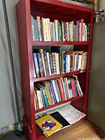 View of vintage red wooden bookshelf with three shelves filled with various vintage books including art books.