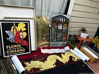 Photo showing two framed art pieces, gold dragon wall toppers laid on black cloth, floral wallpaper roll, and white decorative sheet on a red table outdoors.