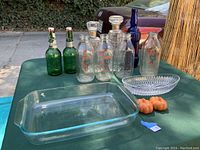 Lot of vintage glassware on a green table including rectangular baking dish, crystal oval bowl, four milk jars with red print, two green glass swing top bottles, and a blue decanter bottle.
