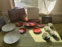 Wide angle photo showing vintage picnic basket, ceramic bowls, red lacquered plates, cream teacups with green saucers, and original boxes on green tablecloth near a window.