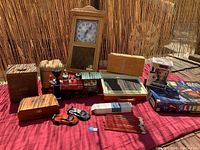 Photo showing all items in the lot arranged on a red blanket outdoors, including a wooden letter toy train, wooden boxes, wooden clock, Eleven Pop doll, Star Wars game, and pencils.