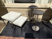 Overall view showing two white square nesting tables and three round tall side tables on a black mat outside.