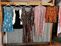 Five vintage women's dresses and tops including floral, printed patterns, and a lace-trimmed pink robe, arranged on a wooden clothing rack.