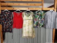 Five women's tops hanging on white plastic hangers on a wooden rack, showing various colors and styles including floral prints, solid red, and pinstriped gray.