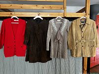 Four vintage women's clothing items hanging on wooden rack: red knit cardigan, dark brown suede coat, beige knit dress with lace and scarf, tan suede belted jacket.
