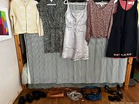 Wide shot of five women's clothing items hanging, including a knit cardigan, dresses, and blouses, with shoes and handbags laid out beneath them.