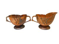 Front and side view of copper creamer and sugar bowl showing Art Deco styled ridges and floral motifs, both with handles and sitting on round bases.