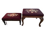 Photo showing two antique needlepoint footstools side by side with floral designs on burgundy fabric seats and wooden legs; smaller has loose trim.
