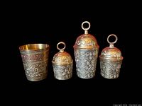 Four items: three brass bells with loop handles and one brass shot glass, all with intricate floral engravings on a black background.