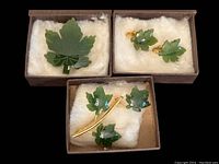 Top-down view of all jewelry pieces in their boxes on white layered padding showing the large single leaf brooch, triple leaf brooch, and maple leaf earrings.