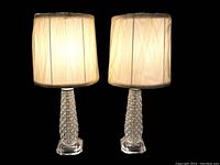 Pair of vintage crystal glass lamps with pleated white fabric shades lit against a black background.