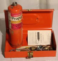 Open orange metal storage case showing propane canister, torch head, and instruction manual inside.