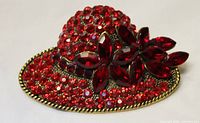 Close-up image showing the red rhinestone brooch pin shaped like a hat with floral motifs, highlighting the various sized rhinestones and gold-tone trim.