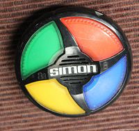 Simon electronic memory game handheld unit placed on a textured surface, showing all four colored pads and the Simon logo in the center.