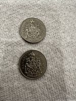 Two Canadian fifty cent coins placed on a textured white surface, showing the reverse side with the Canadian coat of arms.