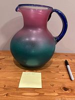 Front side of large Acapulco glass pitcher showing pink to teal gradient and blue handle on wooden surface.