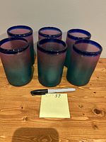 Front view of all six Acapulco glasses showing their size and color gradient from teal to purple with blue rims, placed on a wooden table alongside a marker for scale.
