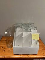 Eight large glass bottles in a white divider box on a wooden surface, one bottle outside the box to display