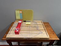 Photo showing the lot items including large Paderno wooden cutting board with juice groove on a wooden table, a small yellow Martha Stewart cutting board propped against the wall, and a rolled red Betty Crocker silicone baking sheet still in packaging on top.