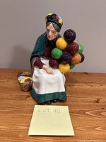 Front angle view of Royal Doulton The Old Balloon Seller figurine seated with balloons and basket.