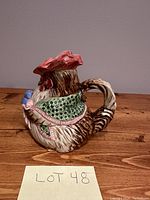 Side view of vintage ceramic creamer shaped and painted as a rooster with a red comb and green patterned body sitting on wooden surface with lot number 48 note visible.