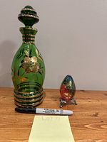 Both the green Murano glass decanter with gold decoration and the multicoloured glass egg paperweight on a metal stand positioned side by side on a wooden surface with a marker for size comparison.
