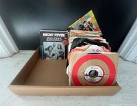 Image showing a large quantity of 7 inch 45 RPM vinyl singles organized in a cardboard box. Visible records and sleeves include Bee Gees 'Night Fever' in a picture sleeve and a Wings single in a red target style sleeve.