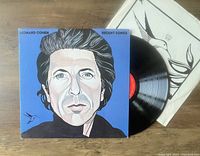 Photo shows the front cover of the Leonard Cohen 'Recent Songs' album with the vinyl partially pulled out along with the inner sleeve showing black and white artwork.