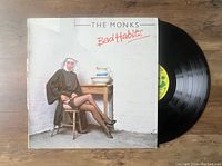 Front cover of The Monks 'Bad Habits' vinyl LP showing a woman in a nun outfit seated on a chair and the vinyl record partially out of the sleeve.