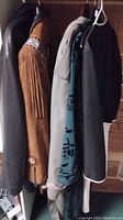 Five hanging garments in closet: black leather jacket, brown suede fringe jacket, beige trench coat, green floral nightgown, black-and-white dress/jacket set