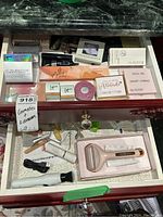 Two drawers shown with various boxed new cosmetics and facial tools including foundations, face palettes, powders, and massage devices.