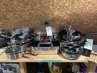Various pots, pans, electric pot, and kitchen accessories on display shelf showing roaster, frying pans, and egg poacher inserts.